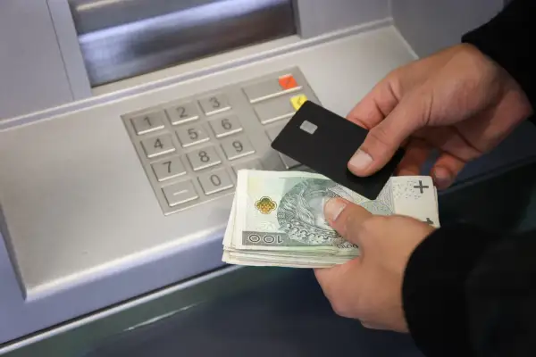 Ręce trzymające banknoty i kartę zbliżają się do wpłatomatu. Pieniądze na koncie czekają na wpłatę.