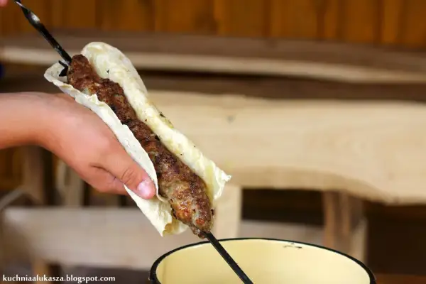 Ręka trzyma lula kebab zawinięty w placek. Mięso jest soczyste i rumiane, gotowe do jedzenia.