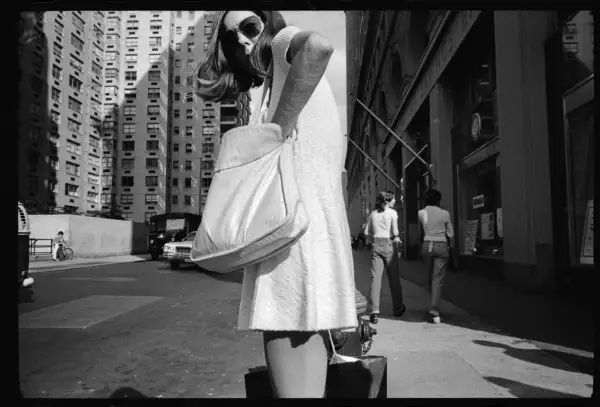 Una donna con borsa a tracolla cammina per strada, con palazzi alti sullo sfondo. Mark Cohen photography cattura la vita urbana.