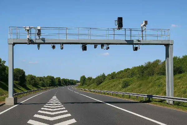 Portique de péage sans barrière sur une autoroute bordée d'arbres. Des caméras et des feux sont installés sur la structure métallique.