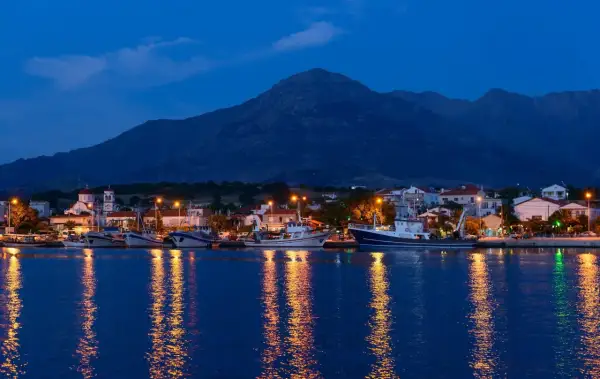 Port z samotraki nocą. Łodzie cumują przy nabrzeżu, a światła odbijają się w spokojnej wodzie. W tle widać góry.