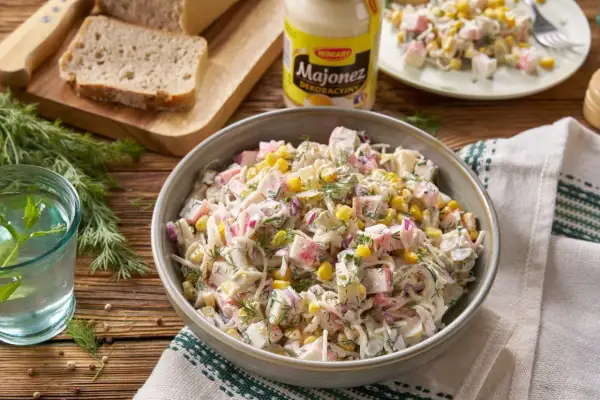 Pyszna sałatka z surimi i ananasem, z kukurydzą i koprem, podana w misce. Obok chleb, majonez i szklanka wody.