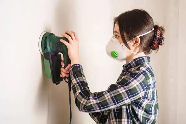 Mujer con mascarilla lijando paredes con una lijadora eléctrica.