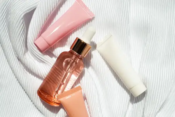 Un frasco de serum con gotero y tubos de crema rosa, blanco y melocotón sobre tela blanca. ¿Que va primero el serum o la crema?