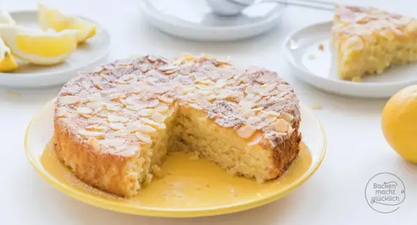 Saftiger Kuchen ohne Mehl, bestreut mit Mandelblättchen und Puderzucker. Dazu Zitronenspalten und eine ganze Zitrone.