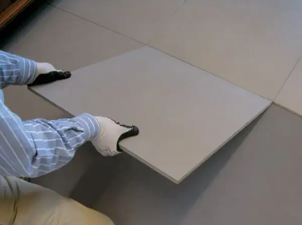 Mains gantées plaçant une nouvelle dalle pour la coller sur du carrelage existant.