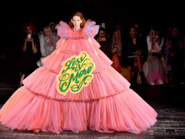 A model wears a voluminous pink tiered gown with a "Less is More" sign, embodying a bold camp aesthetic on the runway.