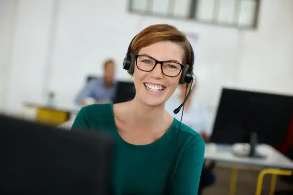 Uśmiechnięta kobieta w okularach i zestawie słuchawkowym, korzystająca z technologii VoIP w biurze.