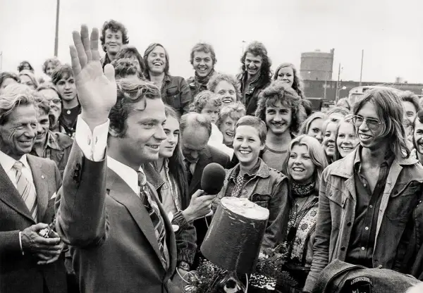 Kung Carl XVI Gustaf vinkar glatt till en folkmassa. En ung kvinna håller fram en mikrofon, kanske för att intervjua en av kungens barn eller någon annan i den glada gruppen.