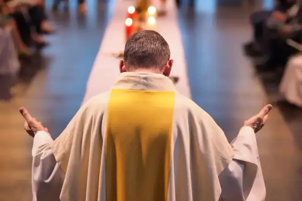 Kapłan w białej szacie z żółtym pasem, z rękami uniesionymi w geście modlitwy, zastanawia się, czy praca w niedziele i święta jest zawsze grzechem. Dylemat zawodowy.