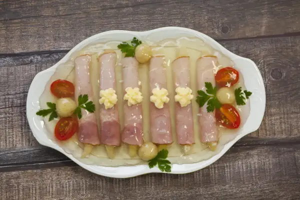 Szparagi w szynce w galarecie, ozdobione pomidorkami koktajlowymi i majonezem, prezentują się apetycznie na białej paterze.