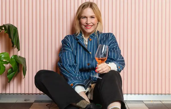 Frida Lund blogg: En glad kvinna med ett glas rosévin sitter på golvet framför en rosa vägg.