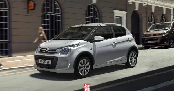 Srebrny Citroën C1 z czarnymi nakładkami na błotniki przemierza miejską ulicę.