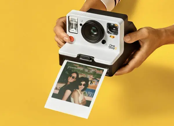 Aparat Polaroid One Step 2 drukuje zdjęcie dwóch kobiet w okularach przeciwsłonecznych na tle kolorowego krajobrazu.