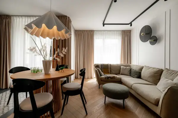 Przytulny salon z beżową kanapą i okrągłym stołem. Nad kanapą w salonie wiszą dekoracyjne lampy, a obok stoi zielona pufa.