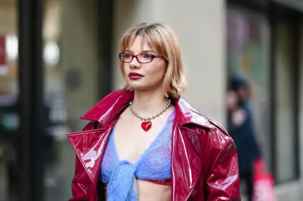 Frau mit roten Brillen und einem stufigen Bob trägt eine glänzende rote Jacke über einem blauen Strickoberteil mit Herz-Halskette.