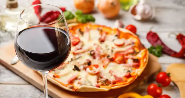 Ein Glas Rotwein neben einer Pizza mit Tomaten und Schinken. Die perfekte Kombination: welcher Wein zu Pizza?