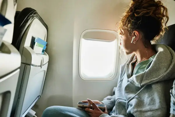 Vrouw met oortjes kijkt uit het vliegtuigraam, hopend op tips wat te doen tegen vliegangst. Ze houdt haar telefoon vast.
