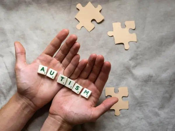 Twee handen houden de letters vast die 'AUTISME' spellen. Puzzelstukjes liggen verspreid, symbolisch voor het spectrum.