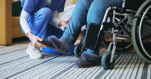 Opiekunka zakłada kapcie osobie na wózku inwalidzkim.
