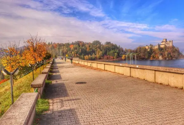 Jesioro Czorsztyńskie jesienią. Spacerowicze i rowerzyści na promenadzie, w tle zamek i kolorowe drzewa.