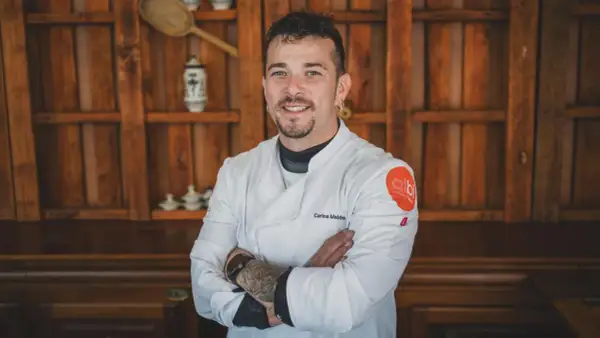 Carlos Maldonado, chef con tatuajes y barba, sonríe con los brazos cruzados frente a una estantería de madera.