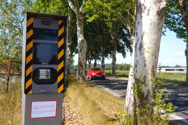 Panneau de **détecteur de radar interdit** sur le bord de la route, avec une voiture rouge passant à proximité.