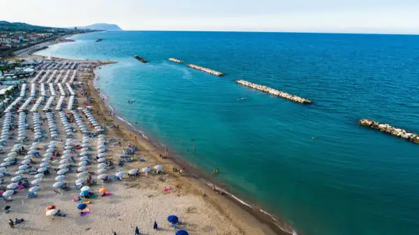 Ampia spiaggia libera a Civitanova Marche, con file ordinate di ombrelloni e lettini, bagnanti che si godono il mare turchese e dighe frangiflutti in pietra.