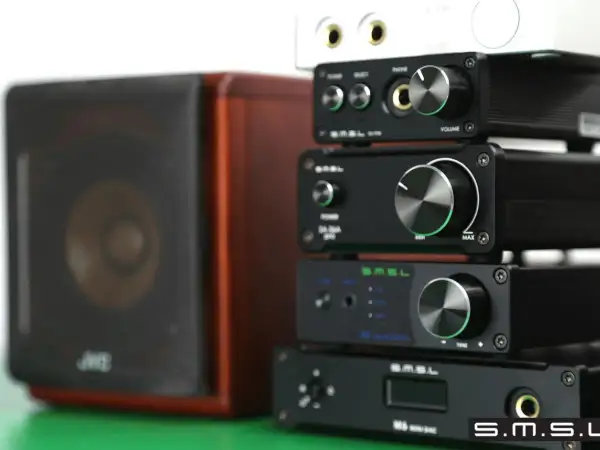 Stosik czarnych urządzeń audio SMSL, w tym SMSL M6 Mini DAC, obok brązowej kolumny głośnikowej.
