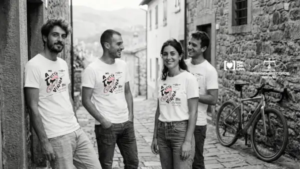 Quattro amici sorridono in un vicolo di pietra, indossando magliette con un disegno di bici e la scritta "A tutta virra". Un'espressione del vocabolario dialetto marchigiano che evoca passione.