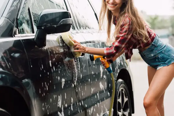 Uśmiechnięta kobieta myje auto gąbką i wężem. Domowe sposoby na czysty samochód to świetna zabawa!