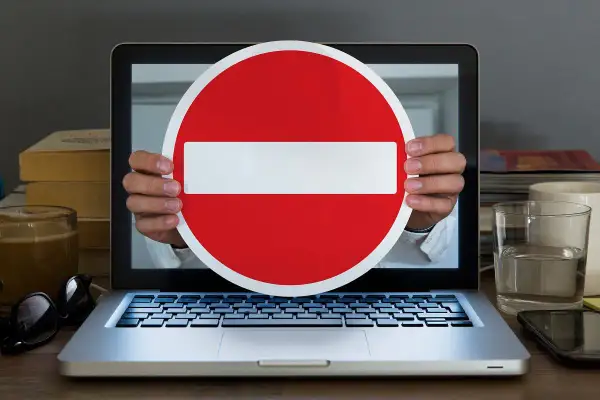 Ręce trzymają znak zakazu wjazdu przed ekranem laptopa, symbolizując cenzurę w internecie.