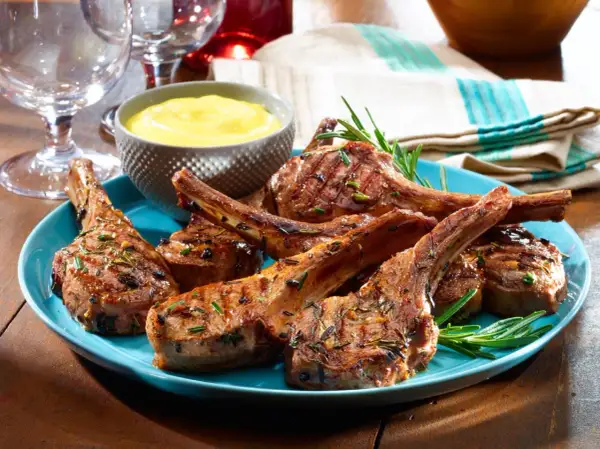 Chuletillas de cordero a la parrilla, jugosas y doradas, servidas en un plato azul con romero fresco y una salsa amarilla.