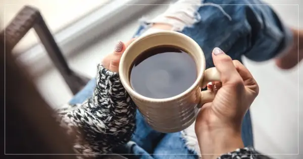 En person med slitna jeans och en stickad tröja håller en mugg med kaffe på morgonen.