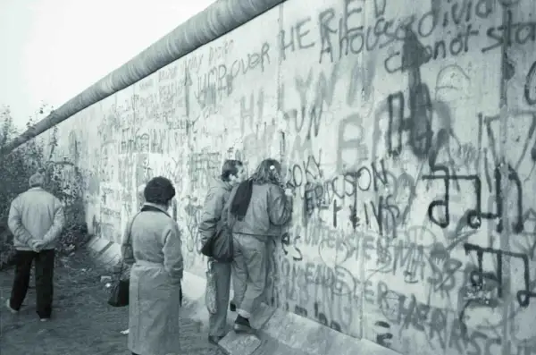 Ludzie oglądają graffiti na Murze Berlińskim, gdy zburzono mur. Widać napisy "Jump over here" i "A house divided cannot stand".