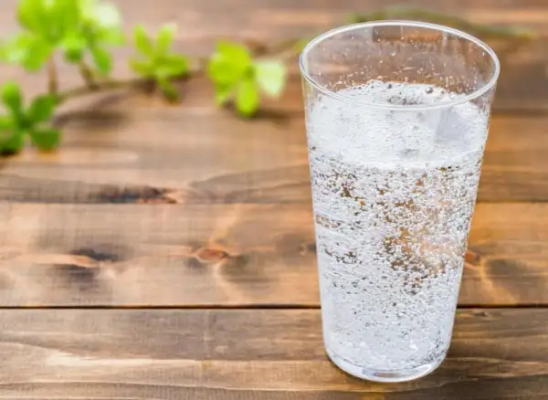 Un verre rempli d'eau gazeuse pétillante, sur une table en bois. Une branche avec des feuilles vertes floues est visible à gauche.
