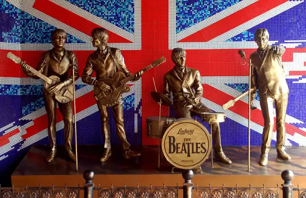 Pomnik The Beatles na tle flagi Wielkiej Brytanii. Czterech muzyków w pozłacanych strojach, z gitarami i perkusją, upamiętnia legendę muzyki.