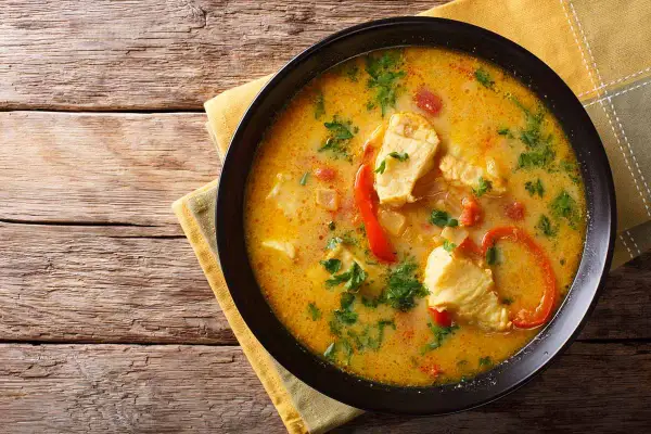Bol de blanquette de poisson crémeuse avec des morceaux de poisson, des poivrons rouges et du persil, servi sur une serviette jaune sur une table en bois.