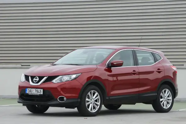 Czerwony Nissan Qashqai J11, z widocznym wykazem żarówek w przednich reflektorach, zaparkowany na tle falistej ściany.