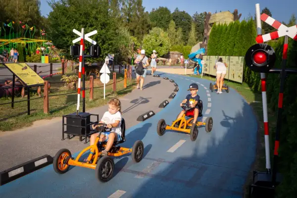 Dzieci ścigają się gokartami na torze w parku rozrywki. W tle widać inne atrakcje i spacerujących ludzi.
