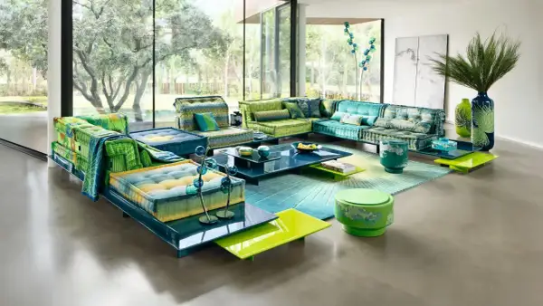 Un salón con sofás modulares en tonos verdes y azules, alfombra degradada y mesas de centro de cristal. Un look boho chic verano perfecto para relajarse.