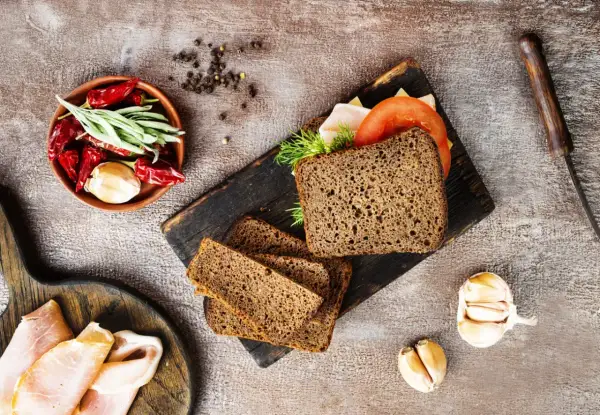 Pyszny chleb z razowej mąki pszennej, idealny na kanapki. Obok składniki: pomidor, ser, wędlina, czosnek, papryczki chili i zioła.
