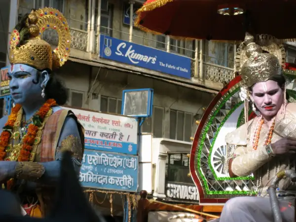 Två män i gyllene krön och smink, en med blå hud, deltar i en procession. Frågan "hur många gudar har hinduismen" kan väckas av deras utseende.