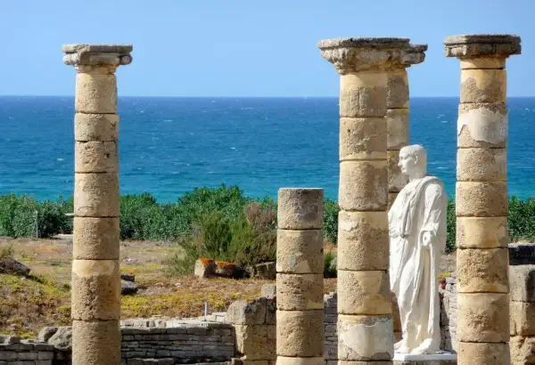 Ruinas romanas en la playa Baelo Claudia, con columnas y una estatua de mármol frente al mar azul.