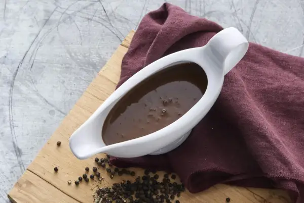 En vit såsslev fylld med mörk brunsås, bredvid en hög med svartpepparkorn på en träskärbräda. Perfekt för ditt brunsås recept.