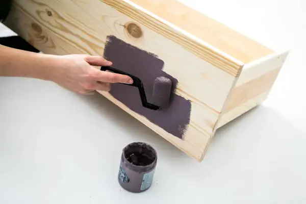 En hand använder en liten roller för att måla en träkista med mörklila färg, vilket ger en jämn yta utan synliga penseldrag.