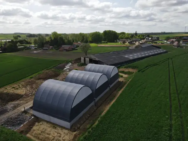 Tre tunnel stalla grigi, moderni, affiancati in un campo verde, con un grande edificio agricolo sullo sfondo.
