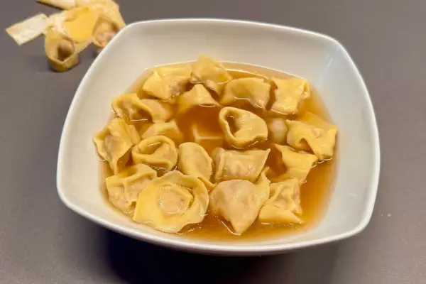 Un piatto fumante di tortellini in brodo, un classico confortante. Sullo sfondo, alcuni tortellini crudi e sfoglia.