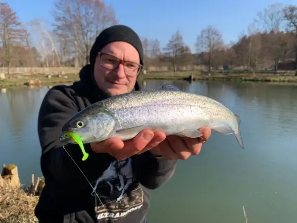 Glücklicher Angler hält frisch gefangene Forelle am Forellenteich. Der Fisch hat einen grellgrünen Köder im Maul.