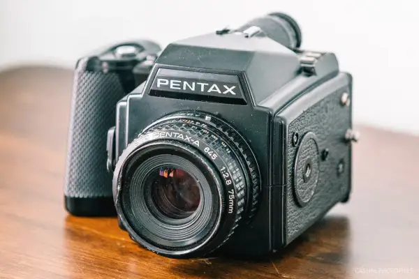 A black Pentax 645 medium format point and shoot camera with a 75mm lens sits on a wooden surface.